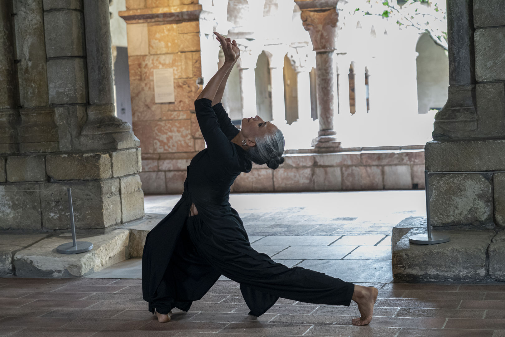 In front of a set of medieval stone pillars, a woman in a flowing navy blue dress strikes a dance pose. She stretches her left leg behind her, her arms and hands raised to the ceiling with palms facing up.